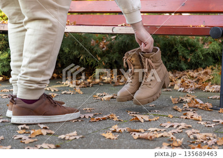 Person's hand lifting light brown women's lace-up boots near comfortable men's sneakers on a concrete path, surrounded by vibrant autumn leaves, illustrating fall fashion and seasonal changes Person's hand lifting light brown women's lace-up boots near comfortable men's sneakers on a concrete path, surrounded by vibrant autumn leaves, illustrating fall fashion and seasonal changes 135689463