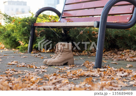 Beige lace-up boots with chunky soles on concrete path among golden autumn leaves by park bench, reflecting fall urban comfort 135689469
