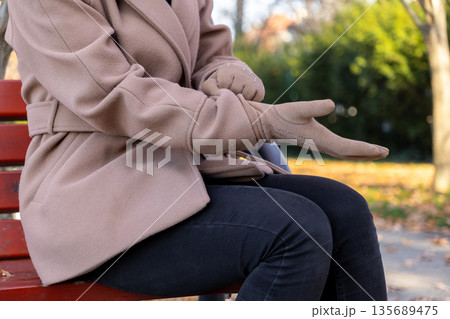 Woman adjusting elegant studded beige glove, seated on vibrant red park bench, symbolizing cozy autumn fashion and comfortable seasonal outdoor style 135689475