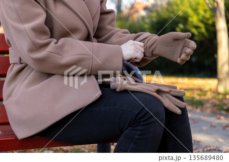 Stylish person in cozy beige coat carefully putting on a warm glove while sitting on a red bench in an autumn park, embracing chilly fall weather 135689480