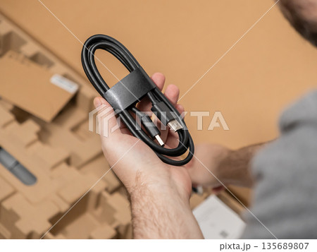 Human hand carefully holding a coiled black USB-A to USB-C charging cable, freshly removed from sustainable cardboard packaging during a new tech product unboxing process Human hand carefully holding a coiled black USB-A to USB-C charging cable, freshly removed from sustainable cardboard packaging during a new tech product unboxing process 135689807