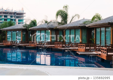 Luxury overwater bungalows with thatched roofs, glass windows and wooden decks by a calm pool. Palm trees, serene tropical vibe. Luxury overwater bungalows with thatched roofs, glass windows and wooden decks by a calm pool. Palm trees, serene tropical vibe. 135692095