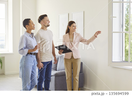 Real estate agent showing empty apartment to family couple buyers, demonstrating nice view from window Real estate agent showing empty apartment to family couple buyers, demonstrating nice view from window 135694599