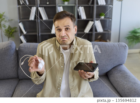 Closeup of man with phone and charger, confused by charging failure at home 135694812