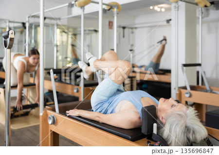 Elderly woman doing lower back exercises while lying on Pilates bed in wellness center Elderly woman doing lower back exercises while lying on Pilates bed in wellness center 135696847