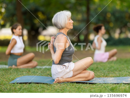 Elderly woman performing Reverse Prayer Gomukhasana during group yoga in park 135696974