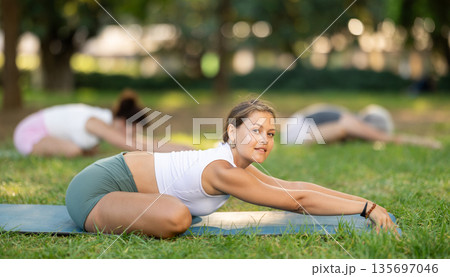 Active happy woman in sportswear sitting on yoga mat and doing stretching legs during yoga class in green park at daytime Active happy woman in sportswear sitting on yoga mat and doing stretching legs during yoga class in green park at daytime 135697046