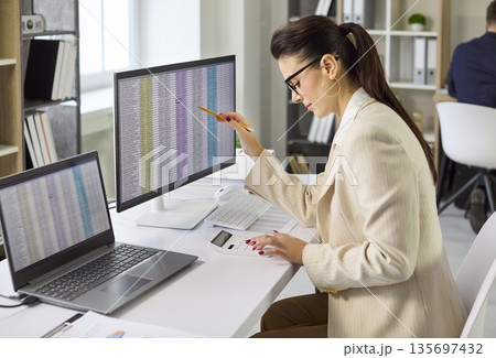 Female accountant analyzing spreadsheets on dual monitors in modern office audit Female accountant analyzing spreadsheets on dual monitors in modern office audit 135697432
