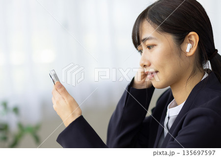 窓辺のテーブルでワイヤレスイヤフォンをつけてスマートフォンを眺めるスーツを着た若い女性 135697597