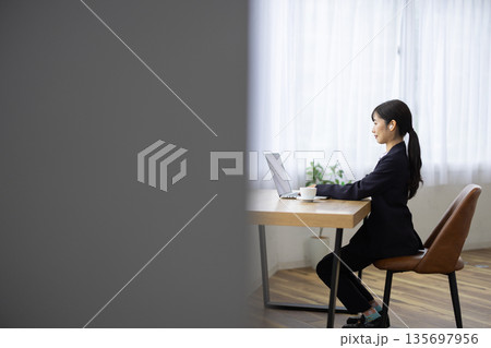 窓際のテーブルでワイヤレスイヤフォンをつけながらパソコン作業をするスーツを着た若い女性 窓際のテーブルでワイヤレスイヤフォンをつけながらパソコン作業をするスーツを着た若い女性 135697956