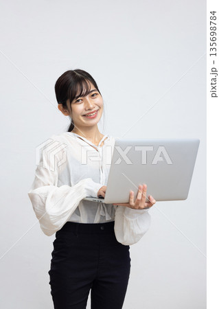 白い背景の前でノートパソコンを操作しながら微笑む若い女性 白い背景の前でノートパソコンを操作しながら微笑む若い女性 135698784