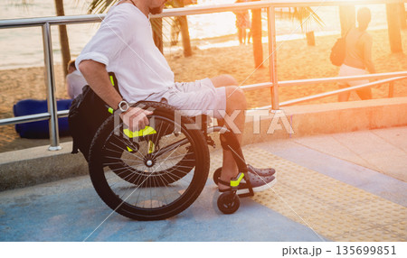 Disabled man in a wheelchair moves on a ramp to the beach. Disabled man in a wheelchair moves on a ramp to the beach. 135699851