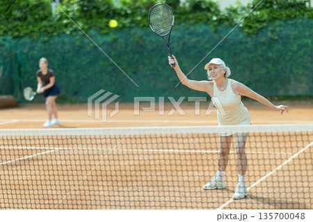 Elderly woman playing doubles tennis on court 135700048