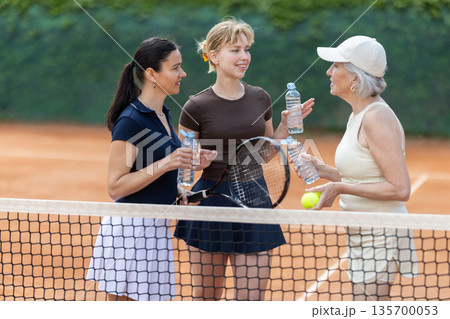 Two girls and senior woman with bottles of water rest during break between tennis games Two girls and senior woman with bottles of water rest during break between tennis games 135700053