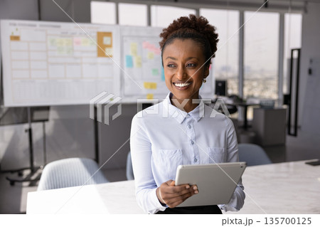 Smiling office employee standing in modern meeting room with tablet 135700125
