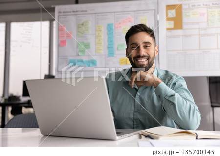 Smiling Hispanic man sits at desk in front of laptop Smiling Hispanic man sits at desk in front of laptop 135700145