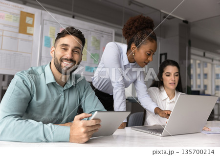 Hispanic man sitting at desk with tablet, two colleagues nearby 135700214
