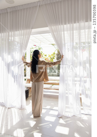 Woman opening white curtains to reveal outdoor space with garden 135700398