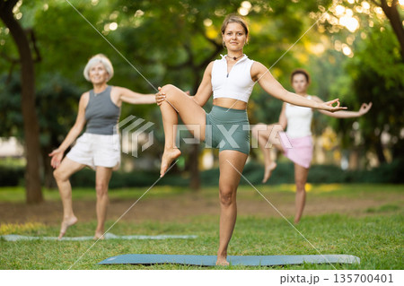 Girl in sportswear exercising with group people in park at daytime, standing in warrior one leg pose on fitness mat Girl in sportswear exercising with group people in park at daytime, standing in warrior one leg pose on fitness mat 135700401