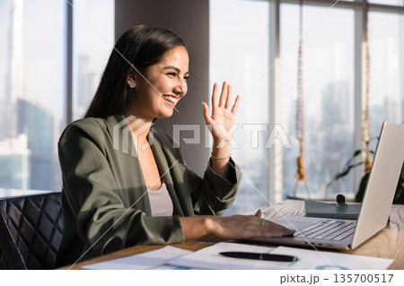 Cheerful Hispanic business professional woman waving hand hello at laptop Cheerful Hispanic business professional woman waving hand hello at laptop 135700517