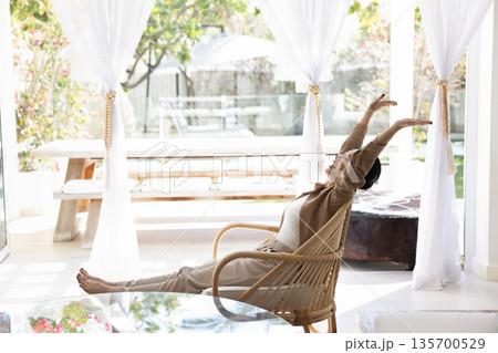 Happy Asian woman sits in wicker chair in outdoor patio Happy Asian woman sits in wicker chair in outdoor patio 135700529