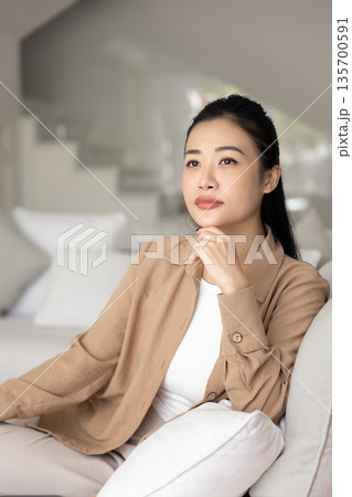 Dreamy Asian woman resting seated on cozy sofa Dreamy Asian woman resting seated on cozy sofa 135700591