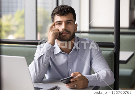 Pensive young man sitting at table contemplating message on cell 135700763