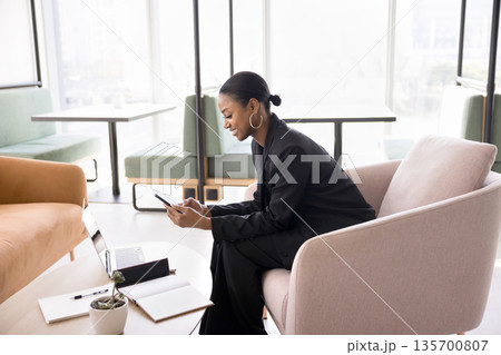 Businesswoman sitting on couch in coworking communicating via phone app Businesswoman sitting on couch in coworking communicating via phone app 135700807
