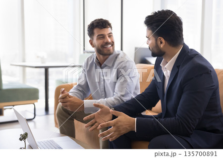 Smiling young businessman talk to male colleague sitting on couch Smiling young businessman talk to male colleague sitting on couch 135700891