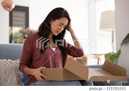 Woman sit on sofa opens cardboard package and feels upset 135701031