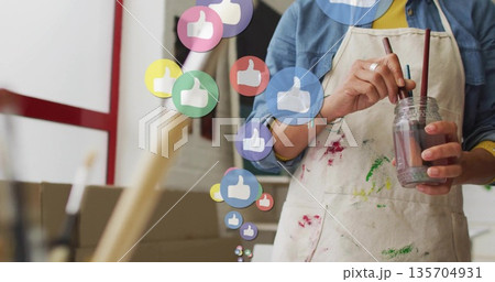 Painter wearing apron mixing brushes in jar at studio, with floating thumbs-up icons, copy space Painter wearing apron mixing brushes in jar at studio, with floating thumbs-up icons, copy space 135704931