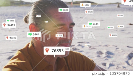 Sitting senior man in collared shirt gazing at horizon on sandy shore, with digital overlay icons 135704970