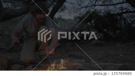 Kneeling man in cargo pants arranging sticks in stone ring in forest clearing, with campfire glow Kneeling man in cargo pants arranging sticks in stone ring in forest clearing, with campfire glow 135704980