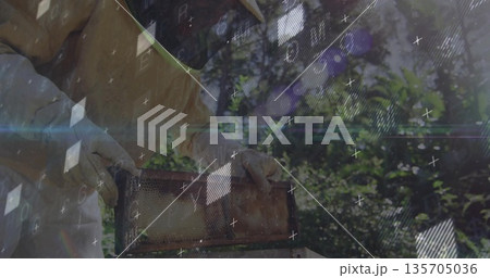 Lifting beekeeper in suit holding wooden honeycomb frame in shaded orchard, with digital overlays Lifting beekeeper in suit holding wooden honeycomb frame in shaded orchard, with digital overlays 135705036