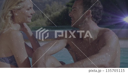 Leaning couple in swimwear chatting at pool edge, with forested hillside background and lens flare 135705115