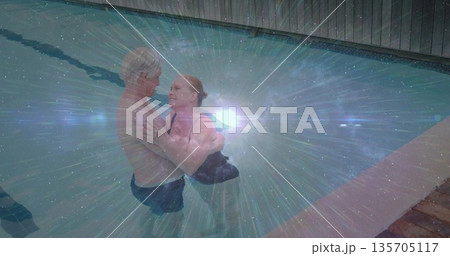 Embracing senior couple standing chest-deep in backyard pool, with dark swimsuits and water glare Embracing senior couple standing chest-deep in backyard pool, with dark swimsuits and water glare 135705117