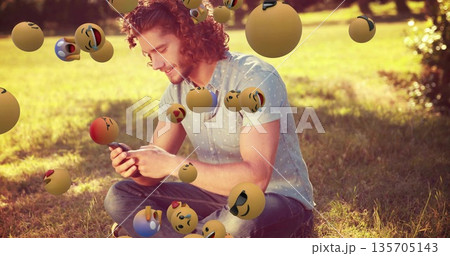 Smiling adult male holding smartphone while sitting on park lawn, with floating emoji spheres 135705143