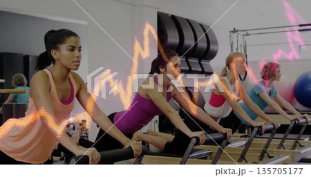 Stretching four women performing leg stretches on Pilates reformer machines in studio, with mirror 135705177