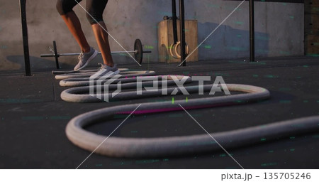 Stepping woman in black leggings and trainers on gym mat, battle rope loops, barbell, copy space Stepping woman in black leggings and trainers on gym mat, battle rope loops, barbell, copy space 135705246