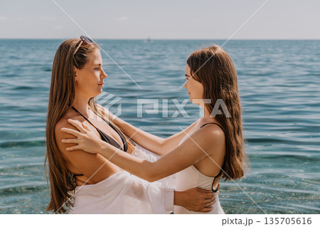 Women beach hugging as they look at each other while standing in the calm blue sea water on a bright summer day Women beach hugging as they look at each other while standing in the calm blue sea water on a bright summer day 135705616