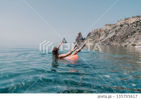 Woman swimming coast relaxes in a bright orange inflatable ring in clear turquoise sea water near rocky cliffs during a summer vacation 135705617