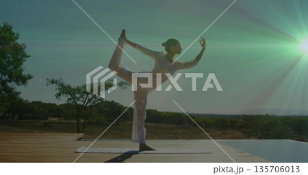 Balancing mid adult woman wearing white outfit in dancer pose on wooden deck, with yoga mat Balancing mid adult woman wearing white outfit in dancer pose on wooden deck, with yoga mat 135706013