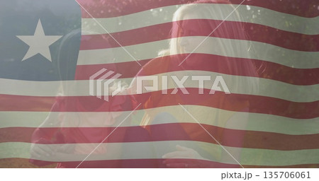 Sitting mother and daughter having picnic on colorful blanket in grassy park, under flag overlay 135706061