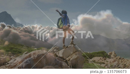 Standing hiker raising arms atop rocky outcrop in mountains, with green backpack and hiking boots 135706073
