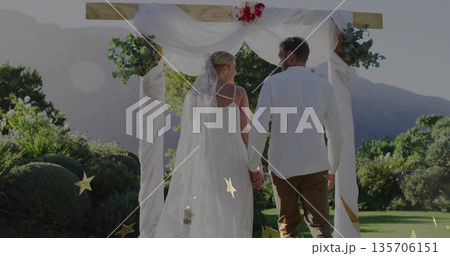 Standing bride and groom holding hands under wedding arch in garden, with drapery, flowers, veil Standing bride and groom holding hands under wedding arch in garden, with drapery, flowers, veil 135706151