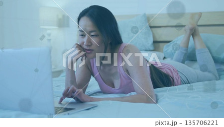 Lying Korean woman in pink top, gray leggings, focusing on laptop in bedroom with network overlay 135706221