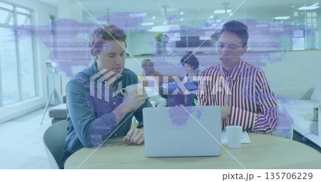 Two colleagues in blue and striped shirts discussing over mugs in open-plan office, with laptop 135706229