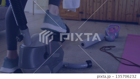 Pedaling woman in dark leggings on stationary bike in home gym, with foam roller and kettlebell 135706232