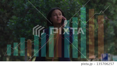 Holding smartphone to ear executive in navy blazer examining digital chart overlays on park walkway Holding smartphone to ear executive in navy blazer examining digital chart overlays on park walkway 135706257