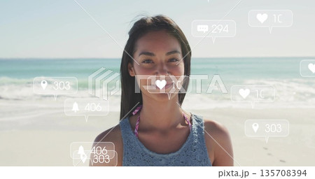 Standing woman wearing blue crochet top over pink bikini on beach, with floating social media icons 135708394
